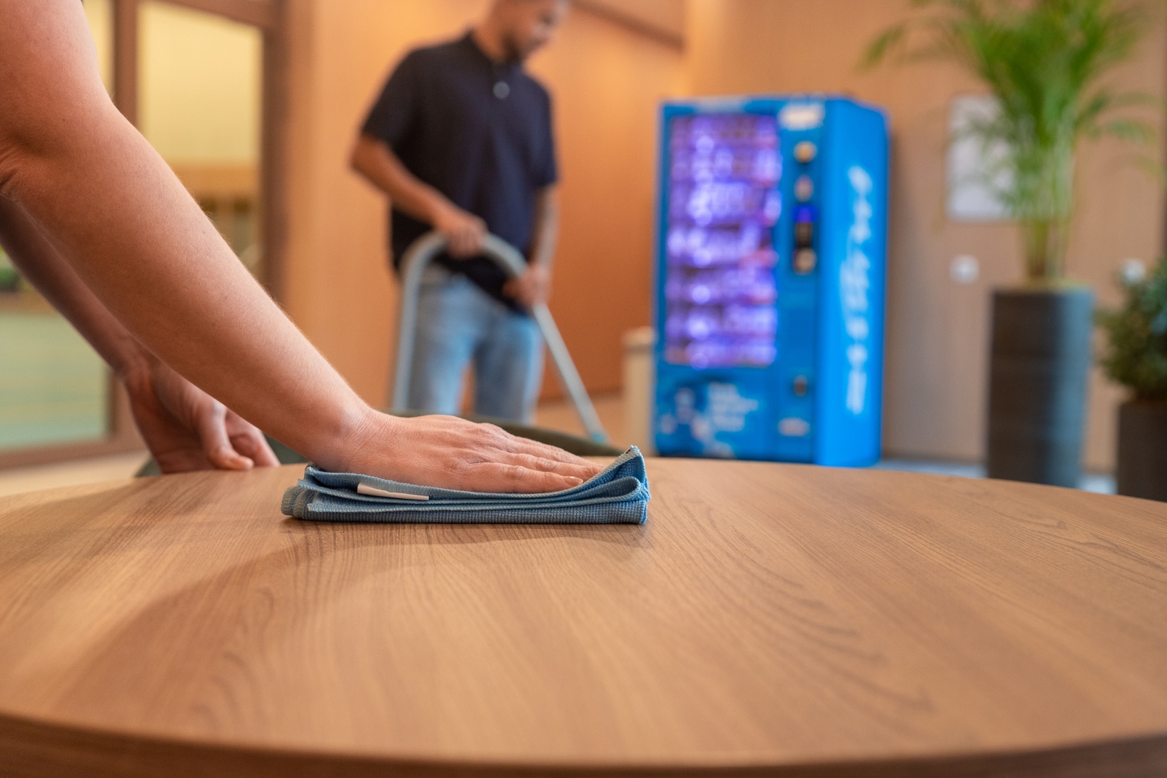 Een schoonmaker gebruikt een microvezeldoek om een tafel schoon te maken, terwijl een andere schoonmaker in de achtergrond stofzuigt